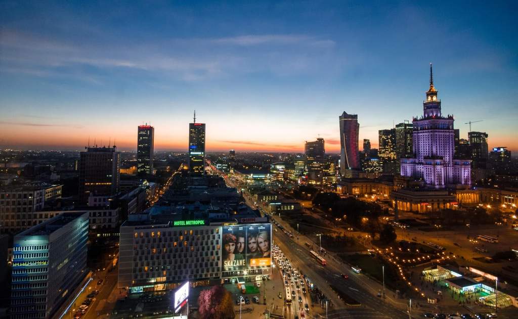 Panoramatický pohled na centrum Varšavy za soumraku. Dominantou je osvětlený Palác kultury a vědy a moderní mrakodrapy, pod nimi rušná křižovatka s auty.