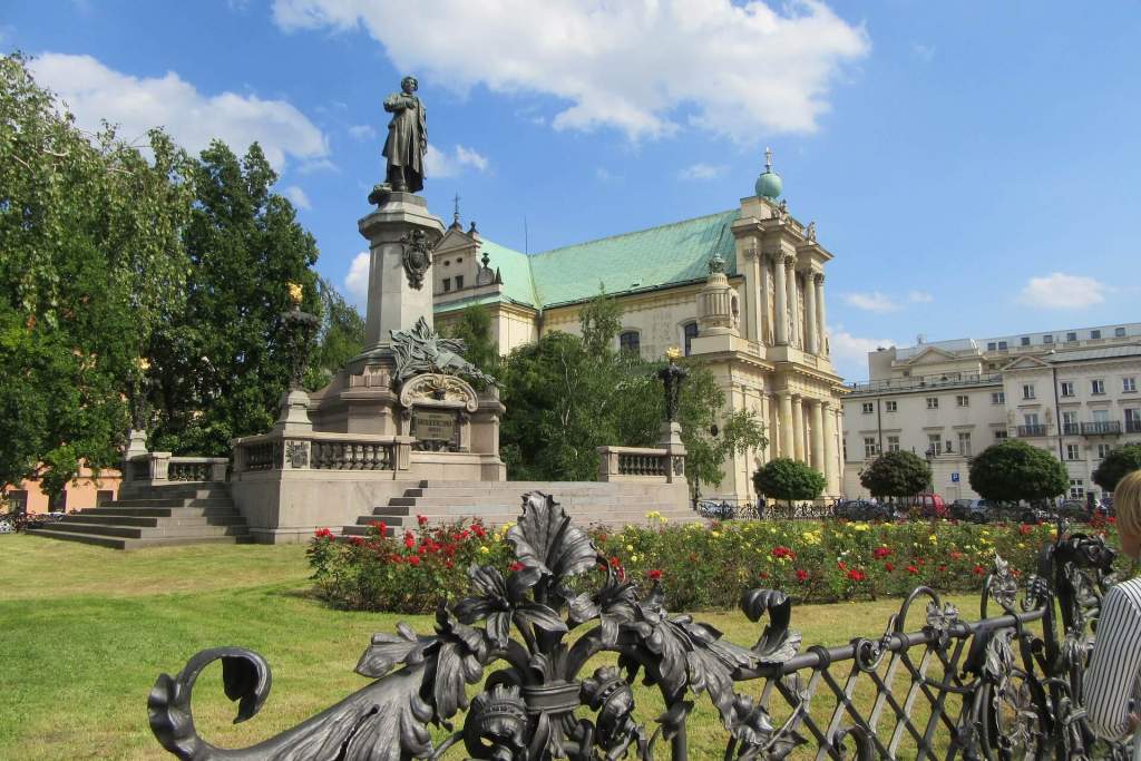 Socha Mikuláše Koperníka a kostel svaté Anny v historickém centru Varšavy za slunečného dne, s růžemi v popředí.