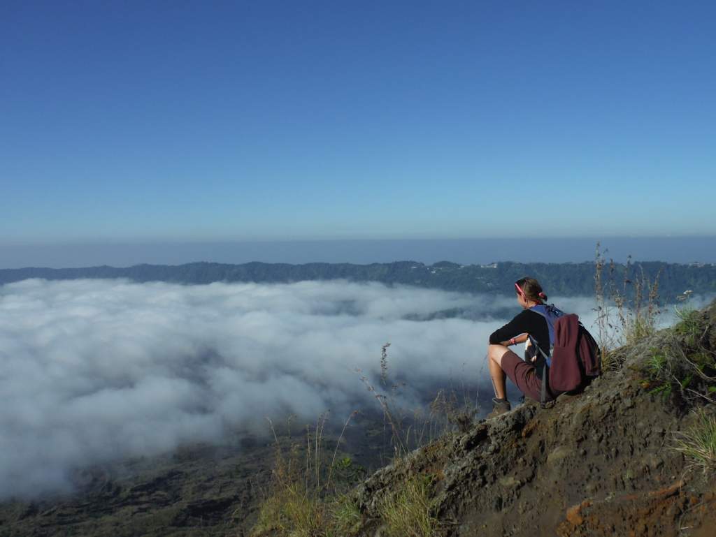 Žena s batohem sedí na skalnatém útesu, shlíží na moře mraků a hory pod jasnou oblohou. Inspirace pro první krok.