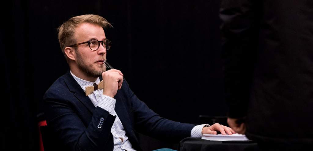 Ladislav Zibura s brýlemi a dřevěným motýlkem, zamyšleně drží pero u úst, před ním poznámkový blok. Portrét z rozhovoru.