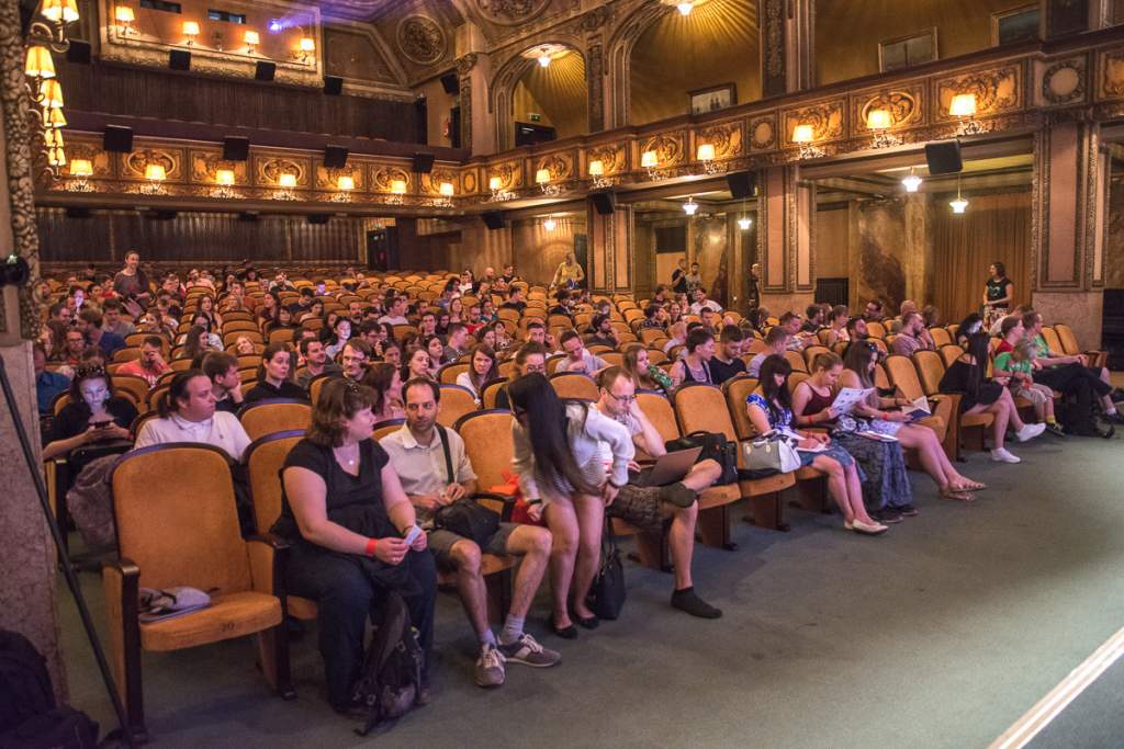 Publikum plného sálu sleduje přednášku během cestovatelské akce. Historický sál s ozdobnými stěnami a sedadly.