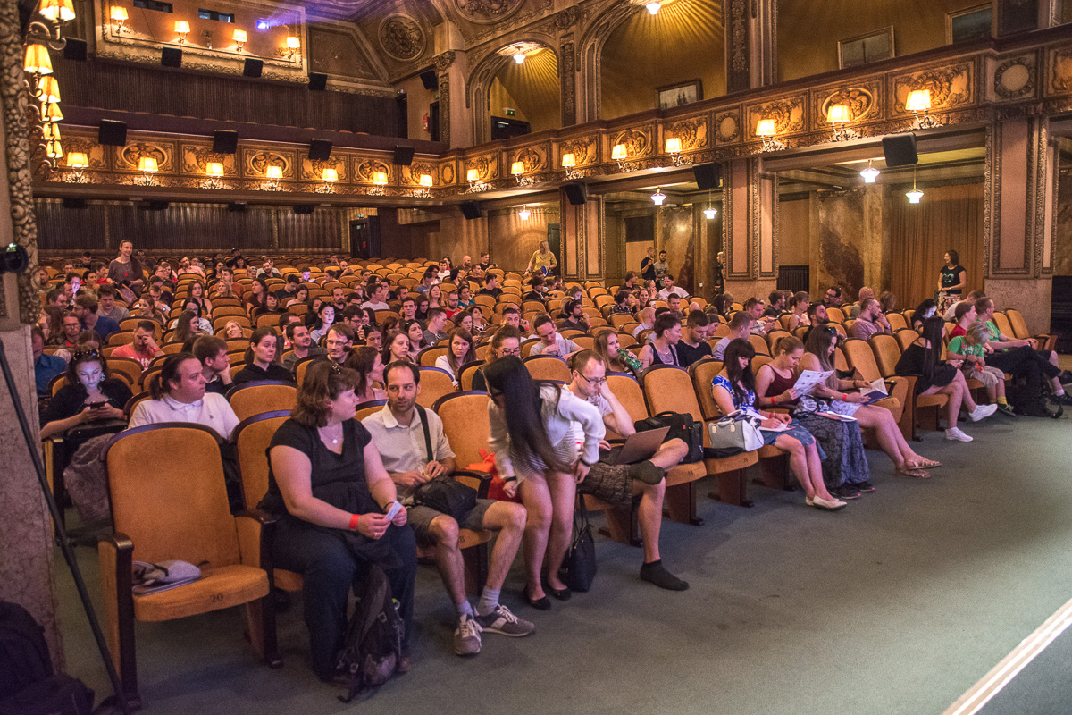 Publikum plného sálu sleduje přednášku během cestovatelské akce. Historický sál s ozdobnými stěnami a sedadly.
