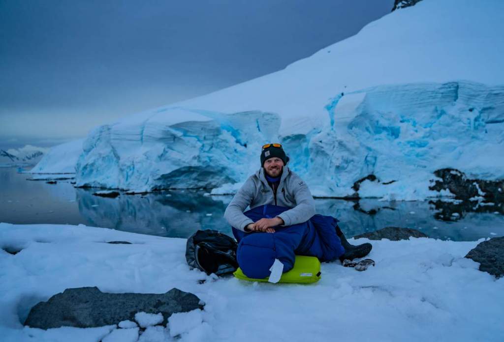 Jan Hvízdal se usmívá a sedí na sněhu s kempingovou výbavou před obrovskými modrými ledovci v Antarktidě.