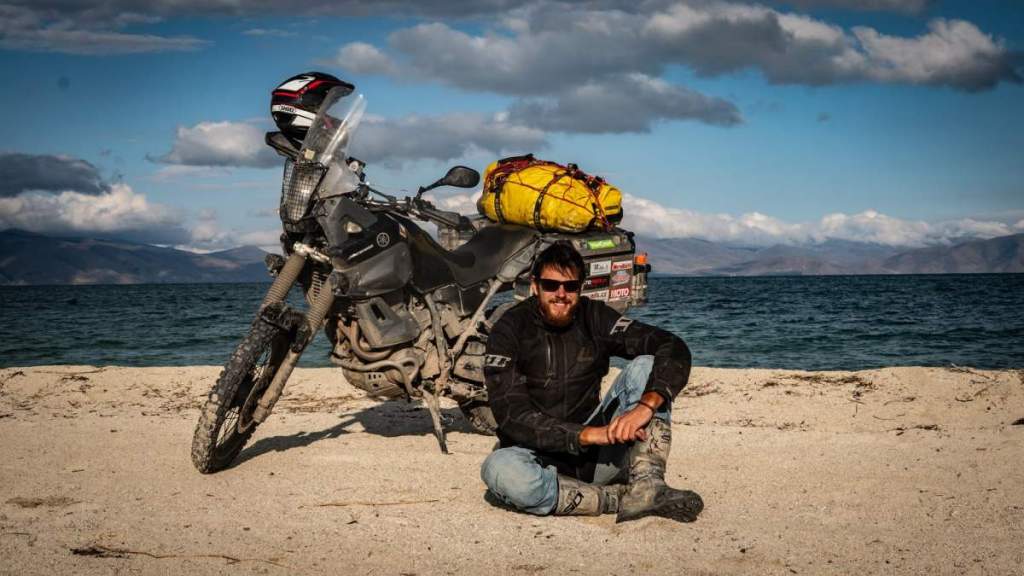 Ondra Vlk sedí na písčitém břehu u velkého jezera vedle cestovní motorky s nákladem. V pozadí se táhnou hory pod částečně zataženou oblohou. Scéna z cesty na Sibiř.