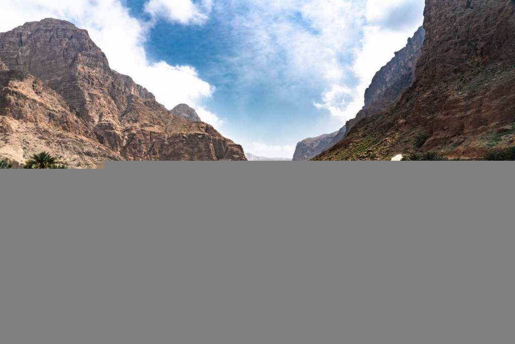 Panoramatický pohled na skalnaté údolí Wadi Al Arabeen v Ománu s vysokými horami, modrou oblohou a palmami v úpatí.
