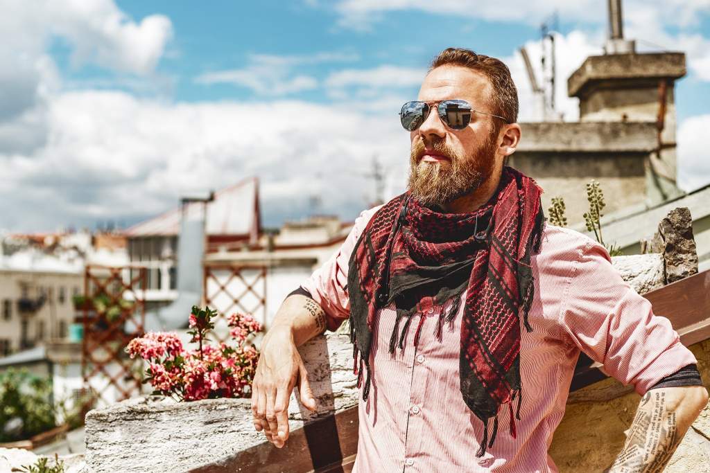 Tomáš Slavíček, digitální nomád, s plnovousem a šálou, opřený o zídku na střeše s výhledem na město a modrou oblohu.