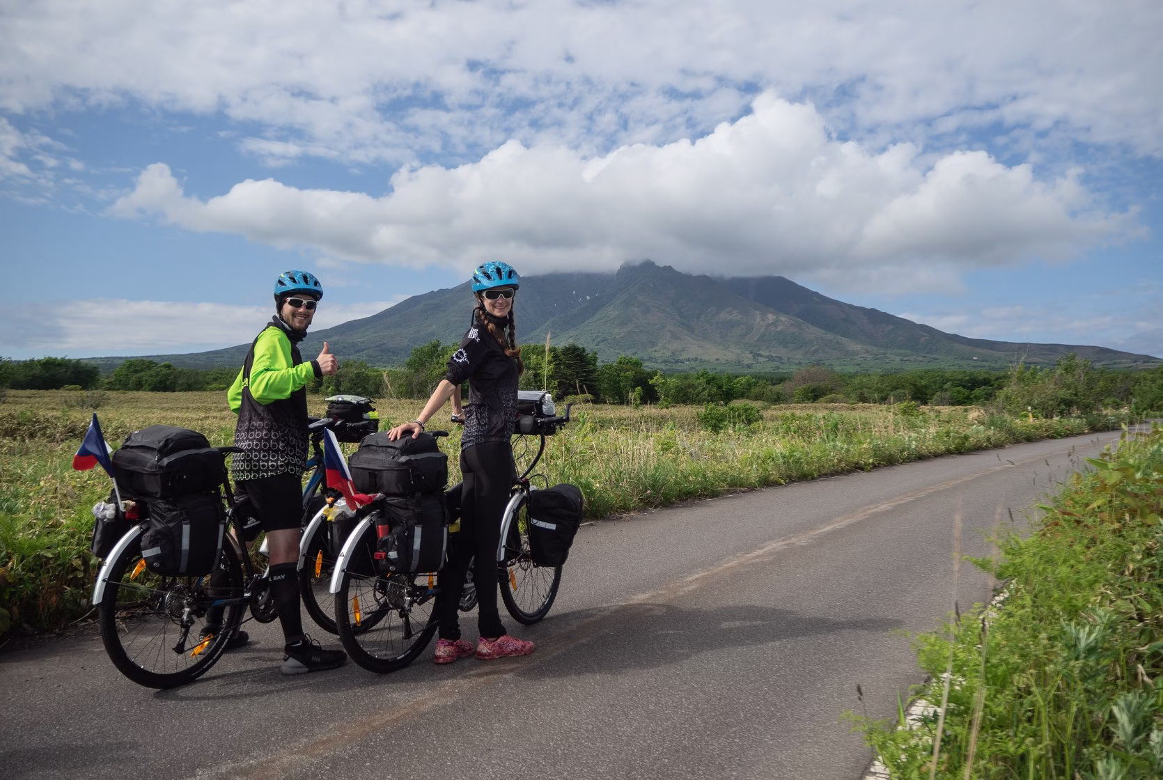 Zuzka Blažková a její partner s plně naloženými cyklistickými koly na cestě v Japonsku, za nimi hora částečně v mracích.