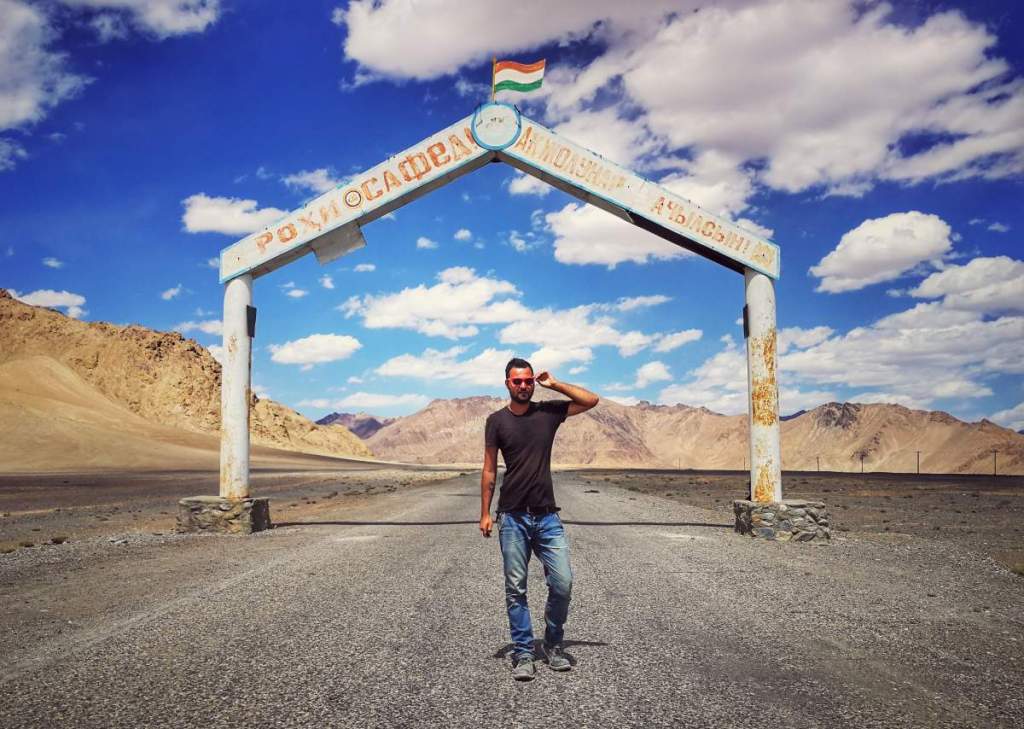 Cestovatel na Pamir Highway v Tádžikistánu, stojí pod vítacím obloukem s tádžickými nápisy a indickou vlajkou. Hory a modrá obloha.