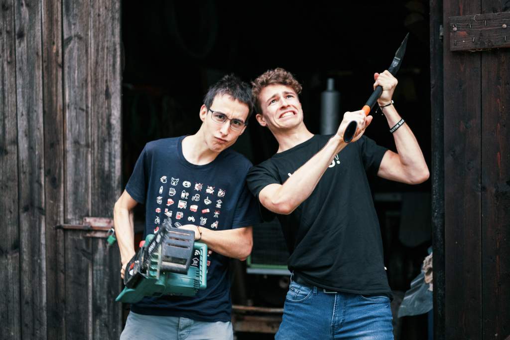 Matouš Vinš s motorovou pilou a kolega s velkou zahradní pilou pózují před dřevěnými dveřmi. Symbolika podnikání a digitálního nomádství.