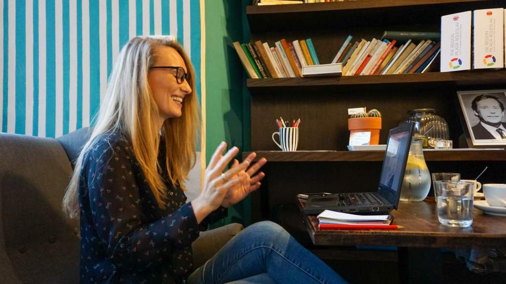 Kika Šrédlová s úsměvem gestikuluje u laptopu v kavárně s knihovnou. Ilustrace k rozhovoru o digitálním nomádství a učení němčiny.