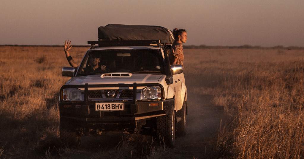 Bílý terénní vůz 4x4 se střešním stanem projíždí africkou savanou při západu slunce. Muž za volantem mává, žena vyhlíží z okna.
