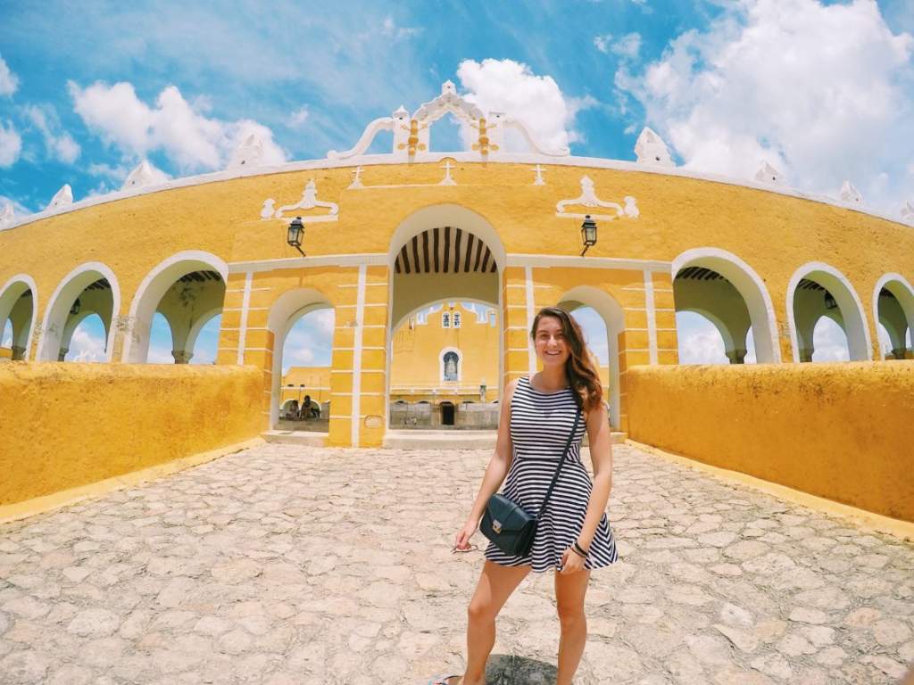 Mladá žena v pruhovaných šatech s úsměvem stojí na dlážděné cestě před žlutým klášterem svatého Antonína z Padovy v Izamalu, Mexiko.