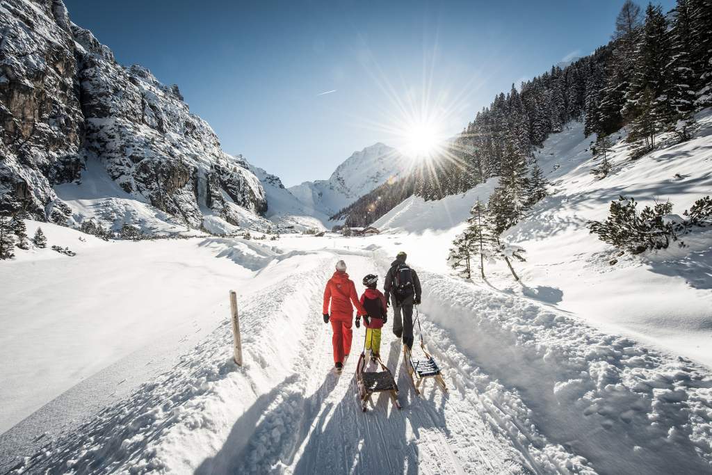 Rodina s dítětem táhne sáňky po zasněžené cestě v údolí Pinnistal v Tyrolsku. Slunečný zimní den v rakouských Alpách, obklopené horami a lesy.