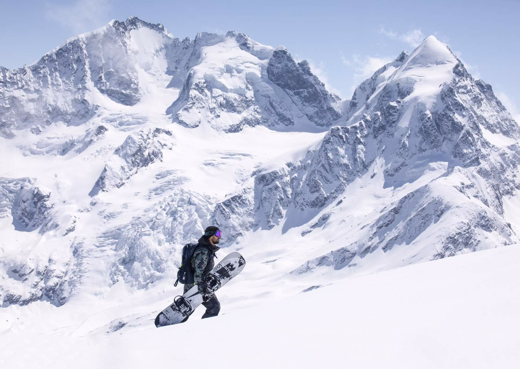 Snowboardista s prknem na zádech stoupá zasněženým svahem, v pozadí majestátní vrcholky Alp pod jasnou zimní oblohou.