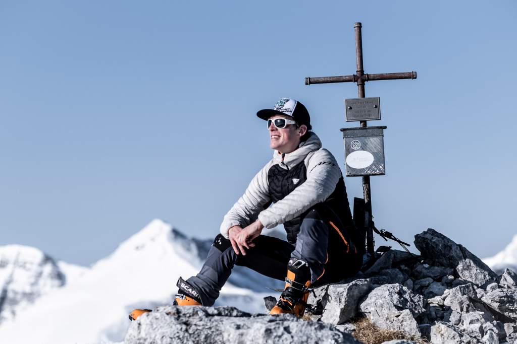 Jakub Cejpek v lyžařském vybavení sedí na vrcholku hory Loigistal v rakouských Alpách u vrcholového kříže, za ním sněhem pokryté štíty.