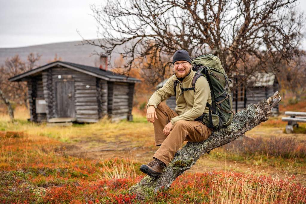 Mladý muž s batohem sedí na kmeni stromu v podzimním Laponsku s červeným vřesem a dřevěnými sruby v pozadí.