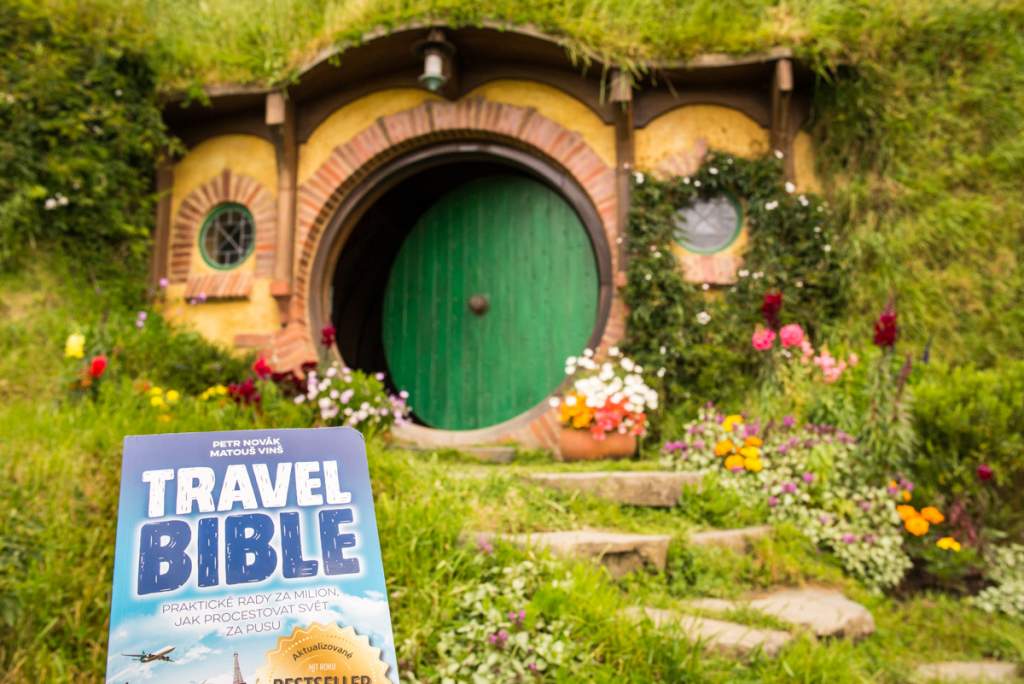 Kniha Travel Bible před kulatými zelenými dveřmi hobitího domu v Hobbitonu na Novém Zélandu, obklopeného květinami a zelení.