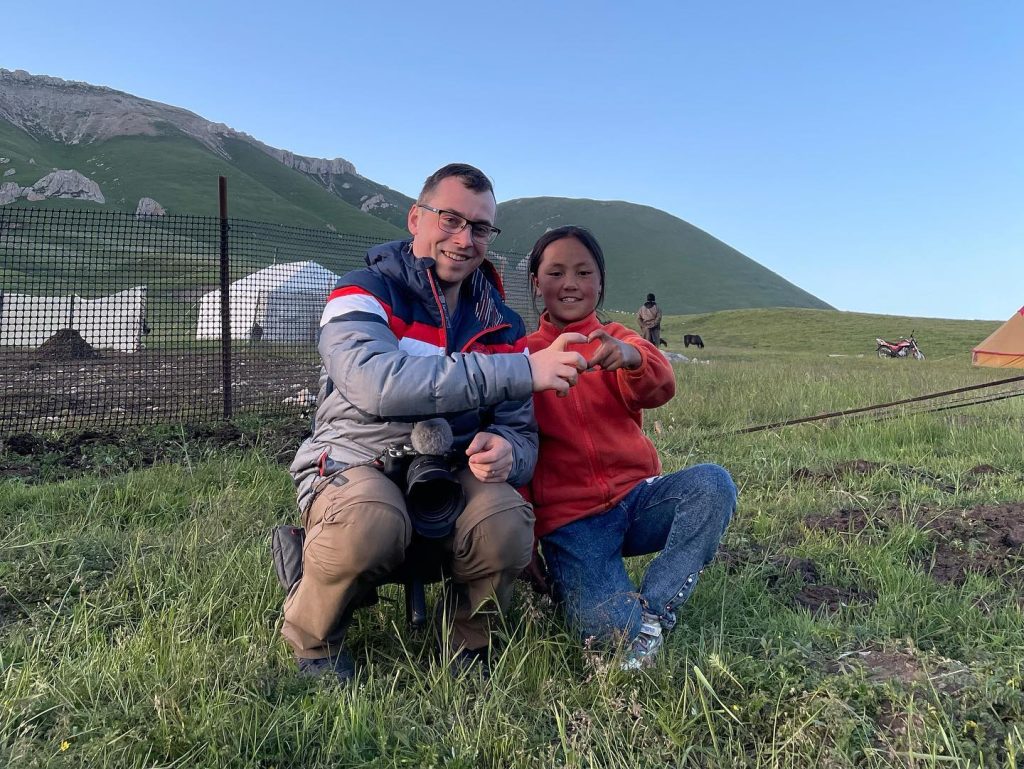 Pavel Dvořák s usměvavou dívkou z čínské vesnice na horské pastvině. Pavel drží kameru, dívka dělá srdce. V pozadí jurty.