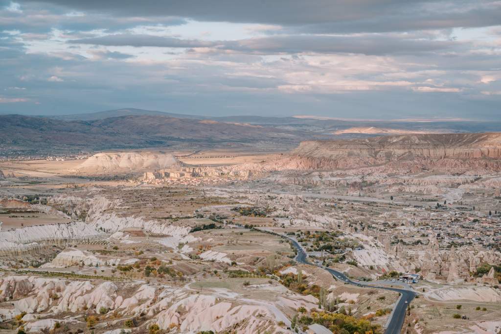 Panoramatický pohled na podzimní Kappadokii s erodovanými údolími, pohádkovými komíny, vesnicemi a klikatou silnicí pod oblačnou oblohou.