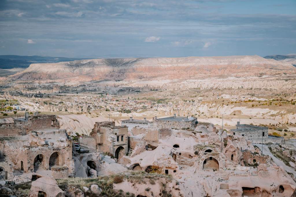 Rozsáhlé údolí s erodovanými skalními útvary a jeskynními obydlími v Kappadokii. V popředí historické domy a hotel, v pozadí pastelově zbarvené kopce pod zamračenou oblohou.