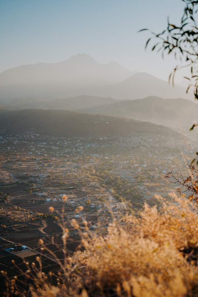 Pohled z výšky na podzimní krajinu Kappadokie se vsí v údolí, zemědělskými poli a mlžnými horami v pozadí za zlatavého světla.