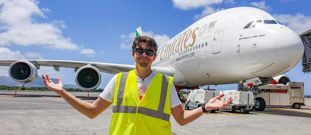 Rosťa Kopecký v reflexní vestě stojí před obrovským letadlem Airbus A380 společnosti Emirates na letištní ploše za slunečného dne.