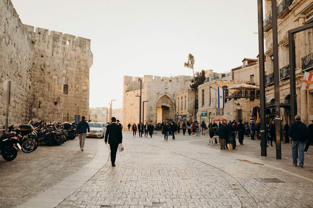 Lidé procházející se po dlážděné ulici před kamennými hradbami a Bránou Jaffa ve Starém Městě Jeruzaléma za jasného dne.