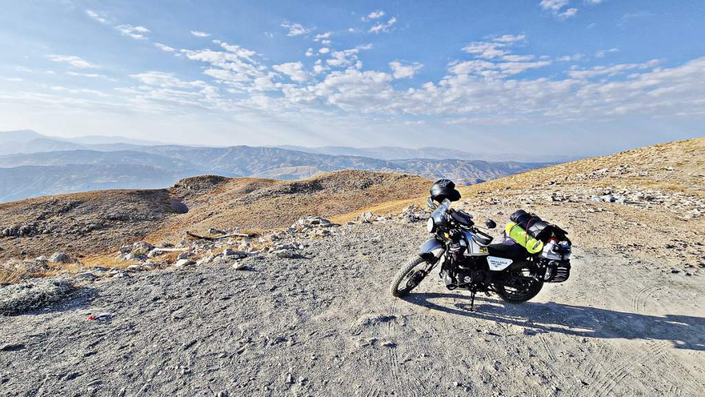 Motorka s cestovní výbavou zaparkovaná na prašné cestě v hornaté krajině Turecka. V pozadí zvlněné hory pod modrou oblohou.