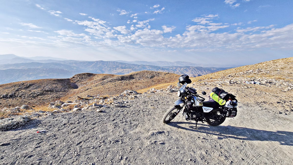 Motorka s cestovní výbavou zaparkovaná na prašné cestě v hornaté krajině Turecka. V pozadí zvlněné hory pod modrou oblohou.
