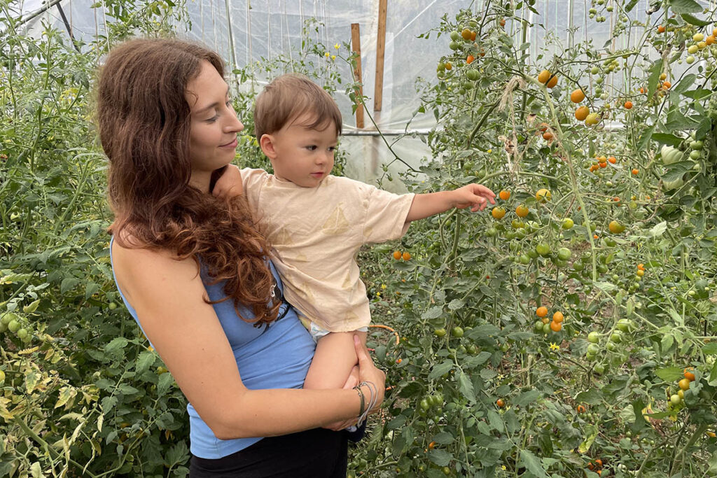Mladá žena drží batole, které ukazuje na zrající rajčata ve fóliovníku. Ukázka rodinného dobrovolnictví na farmě.