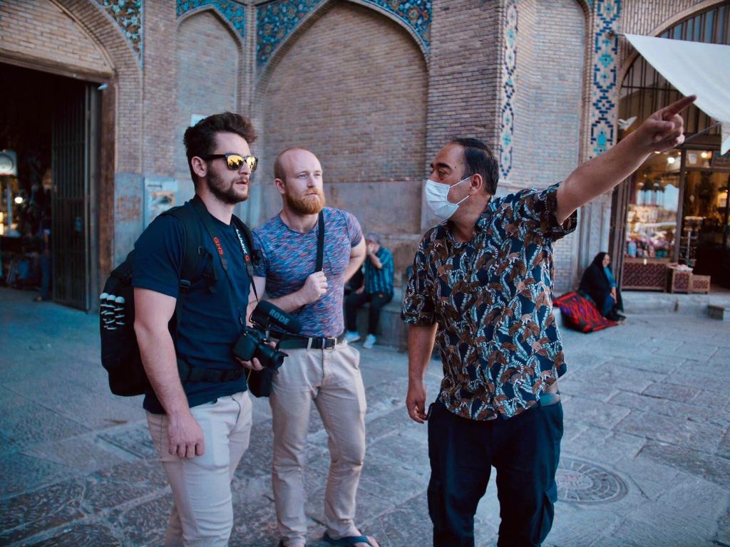 Cestovatelé s kamerou a mikrofonem nahrávají podcast na historickém bazaru. Místní průvodce v masce ukazuje směr.