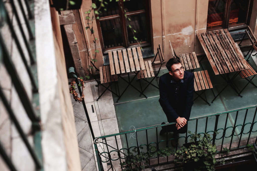 Matouš Vinš z Travel Bible s brýlemi, opřený o tmavé kovové zábradlí balkonu nad venkovní kavárnou.