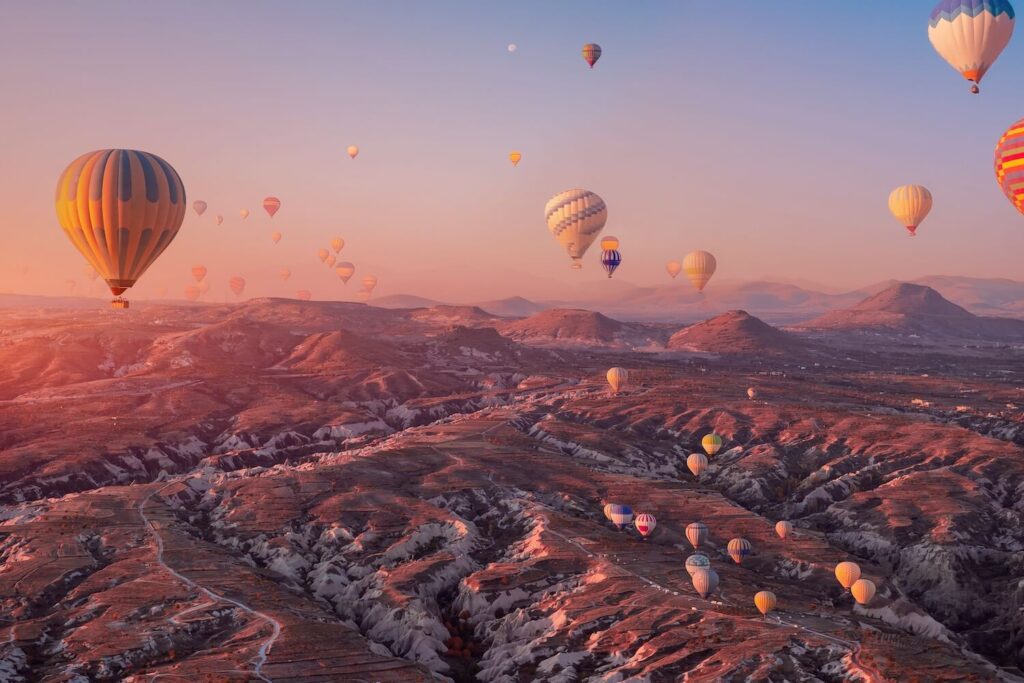 Desítky horkovzdušných balónů nad pohádkovou krajinou Kappadokie v Turecku při východu slunce, obloha v oranžových odstínech.
