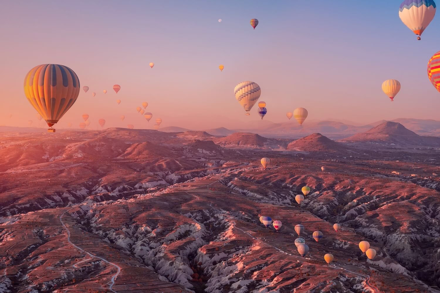 Desítky horkovzdušných balónů nad pohádkovou krajinou Kappadokie v Turecku při východu slunce, obloha v oranžových odstínech.