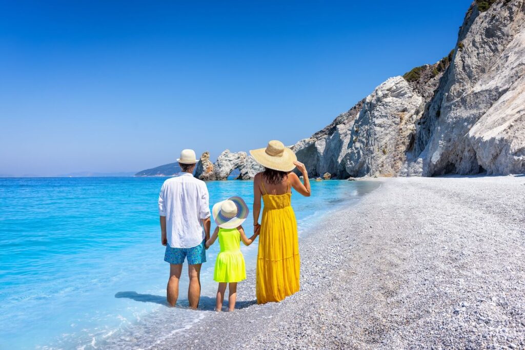 Rodina (rodiče s dítětem) stojí na oblázkové pláži s tyrkysovou vodou a bílými útesy, dívají se na moře. Ideální místo pro last minute dovolenou.