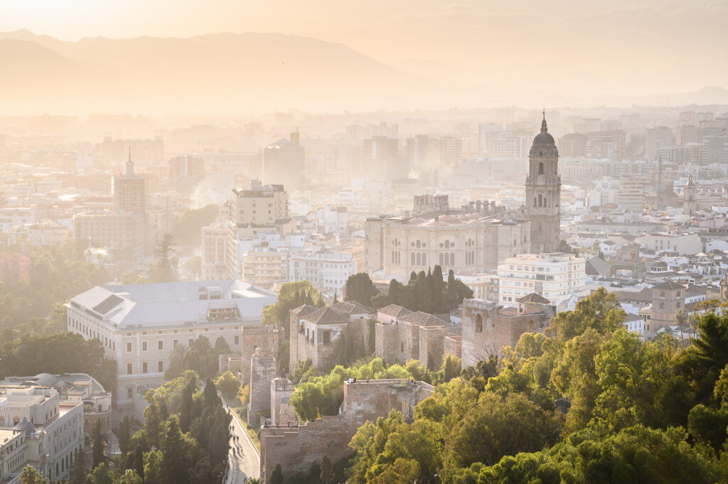 Panoramatický pohled na historické centrum Málagy s pevností Alcazaba a katedrálou, zahalené do podzimního oparu a teplého světla.