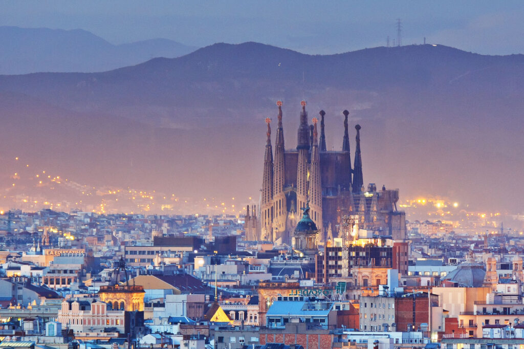 Panoramatický pohled na Barcelonu za soumraku s ikonickou Sagradou Famílií v centru, zářícími světly města a pohořím Collserola.