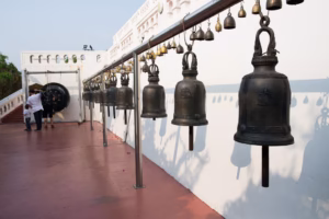 Řada zvonků visících před bílou budovou na hoře Zlatá hora (Wat Saket) v Bangkoku. V pozadí je vidět gong a lidé.