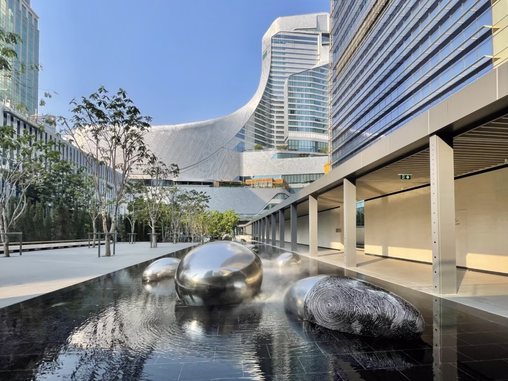 Moderní architektura s vodní plochou a sochami mezi budovami One City Centre a Central Embassy v Bangkoku za slunečného dne.