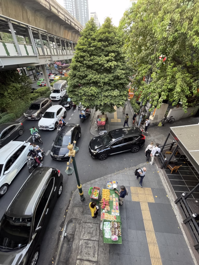 Pohled shora na rušnou ulici v Bangkoku s hustou dopravou aut, motorek a tuk-tuků, Skytrain dráhou nad hlavou a stánkem s ovocem na chodníku.