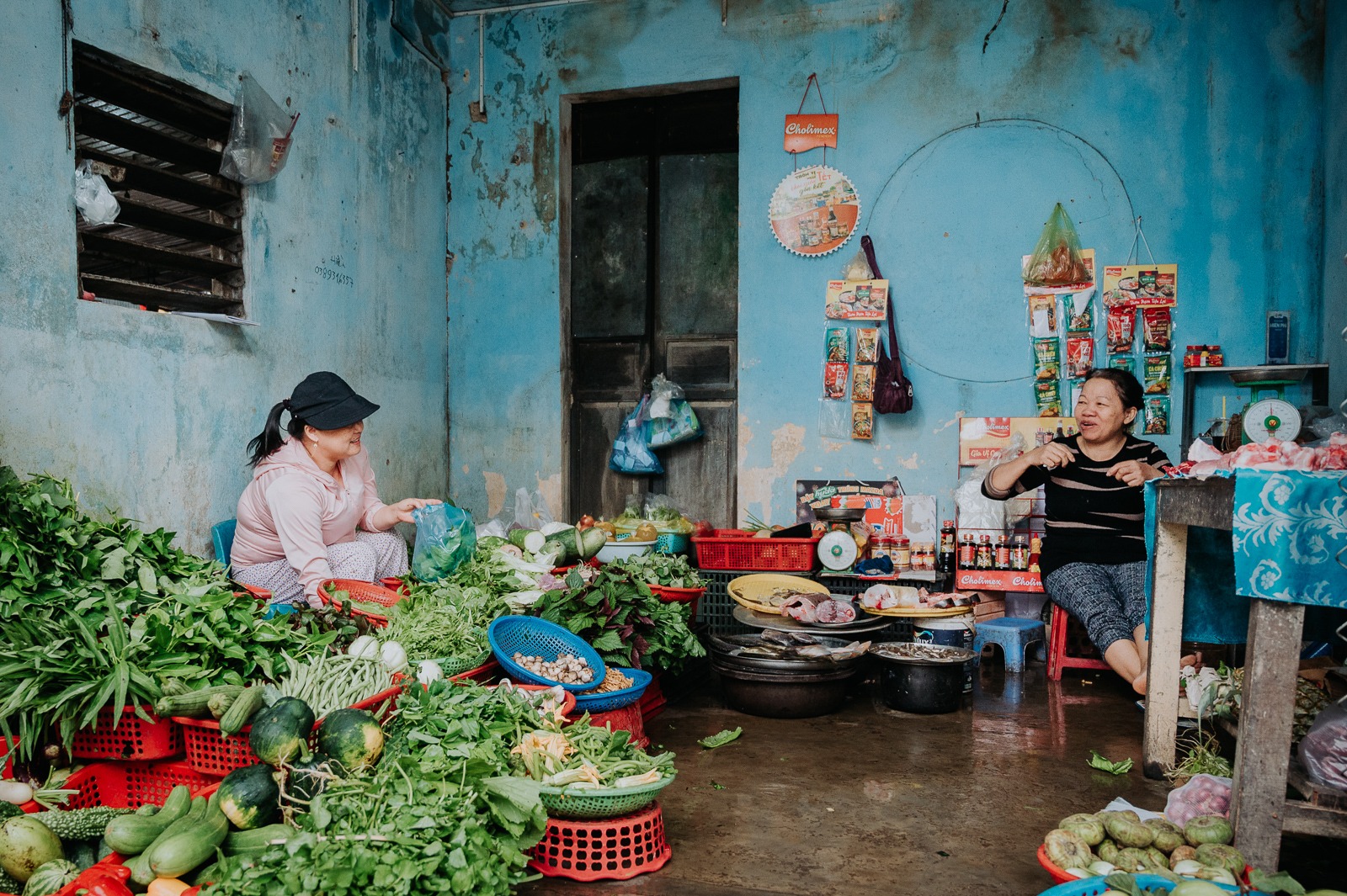 Dvě vietnamské trhovkyně si povídají mezi hromadami zeleniny v modře omítnuté tržnici